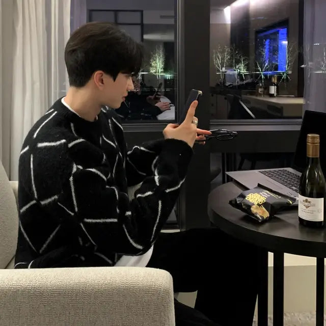 A young man with dark hair sits at a modern round table in a dimly lit, stylishly decorated room. He is wearing a black sweater with a white geometric pattern and holds a smartphone in one hand and eyeglasses in the other. On the table, there is a bottle of wine, a laptop, and some snacks. The large window behind him reveals a nighttime cityscape with illuminated trees and buildings reflecting in the glass. The setting exudes a sophisticated and cozy ambiance.