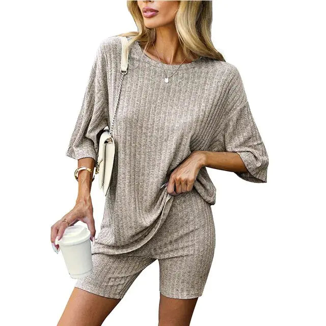 Woman wearing a matching ribbed mauve loungewear set consisting of a loose-fitting oversized top and fitted shorts, accessorized with a crossbody bag, bracelet, and holding a white takeaway coffee cup.