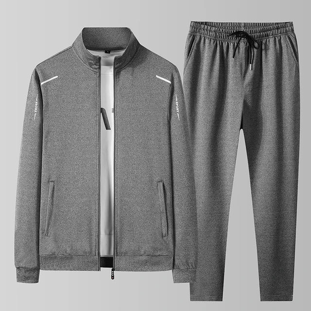 A gray athletic tracksuit consisting of a zip-up jacket and matching jogger pants, displayed against a light gray background. The jacket features a stand-up collar, full-length zipper, two side pockets, and subtle white accents on the shoulders and sleeves. The matching joggers have an elastic waistband with a drawstring, tapered legs, and side pockets. The fabric appears to be a smooth, stretchy material, suitable for sports or casual wear.