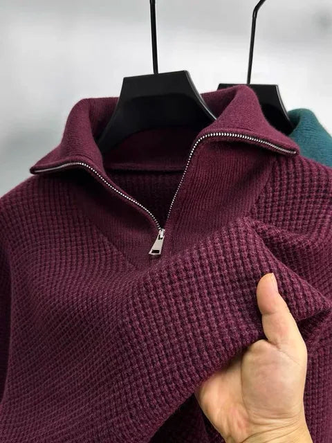This image shows a close-up of a textured, waffle-knit pullover sweater in a rich burgundy color. It features a silver half-zipper with a folded-down collar, giving it a stylish and cozy look. A person is holding up the sleeve to showcase the fabric's softness and texture. In the background, another sweater in a teal shade is visible on a hanger, adding a contrast to the scene.