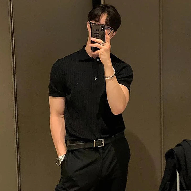 A stylish man with a fit physique takes a mirror selfie in a dimly lit room. He is wearing an all-black outfit consisting of a fitted polo shirt, tailored pants, and a black belt. His left arm is tucked into his pocket, showcasing a silver watch on his wrist. He holds a smartphone in front of his face, partially obscuring his features, while his dark hair is neatly styled. The background features a reflective surface, possibly a wardrobe or mirror, with a dark-colored jacket draped over a chair in the lower