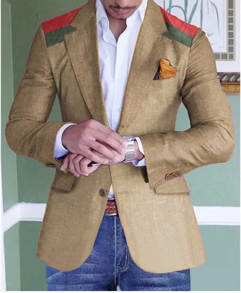 A well-dressed man wearing a tailored tan blazer with red and green shoulder accents, a white dress shirt, and blue jeans. He is adjusting a silver wristwatch on his left wrist. A mustard-colored pocket square with a hint of purple is neatly tucked into the blazer’s breast pocket. The background features a green wall with white trim and a framed artwork