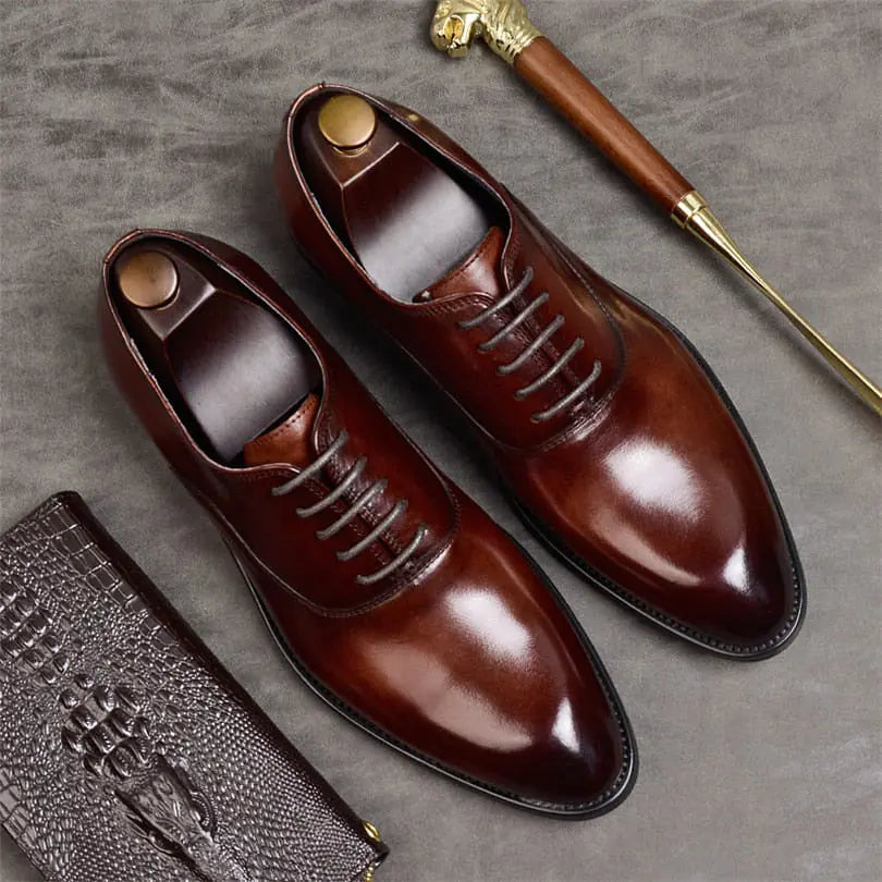A pair of elegant burgundy leather loafers with a glossy finish. The shoes feature gold-toned metal horsebit detailing across the uppers and are displayed with wooden shoe trees inserted. They have a sleek, elongated silhouette with subtle stitching along the toe cap. The loafers are placed on a dark surface, partially framed by a wooden picture frame, emphasizing their classic and sophisticated style.