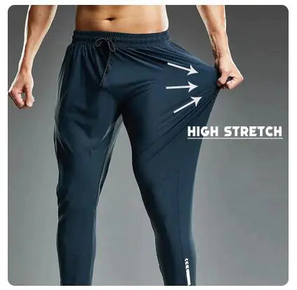 A muscular man wearing dark athletic joggers demonstrates the high-stretch capability of the fabric by pulling the side of the pants outward. The image has a dark gradient background, and bold white text that reads "HIGH STRETCH" with three arrows pointing to the stretched fabric, emphasizing its elasticity. The joggers feature an elastic waistband with a drawstring and a sleek, tapered fit, ideal for sports and activewear.
