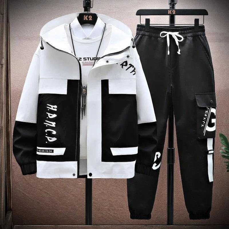 A stylish, modern two-piece streetwear outfit displayed on hangers against a wooden-textured background. The outfit includes a white and gray hooded jacket with a front zipper, multiple pockets, and bold text details on the sleeves and lower part. Underneath the jacket is a white t-shirt with 'K2 Studio' printed on the chest, along with a pendant necklace. The matching black jogger pants feature a white drawstring, cargo-style side pockets, and graphic text accents, giving the ensemble a sporty...