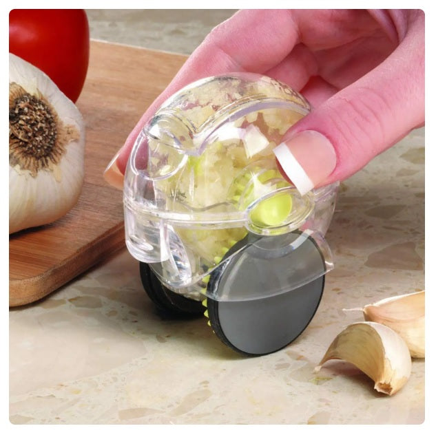 Hand using a rolling garlic chopper with a transparent design and black wheels to mince fresh garlic. Compact and easy-to-use kitchen tool on a marble countertop with garlic cloves and a wooden cutting board in the background.