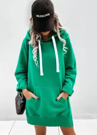 Stylish woman wearing an oversized bright orange hoodie with white drawstrings, paired with white shorts, green and white sneakers, a black Balenciaga baseball cap, and a black quilted handbag, posing against a light grey background.
