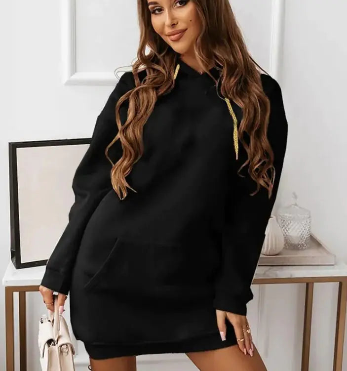 Woman wearing a long-sleeve, oversized maroon hoodie dress with a front kangaroo pocket and gold drawstrings. She is holding a light-colored handbag and posing indoors with a soft smile. The outfit has a casual, cozy look perfect for lounging or casual outings.