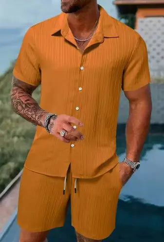 A stylish man with a muscular build is wearing a short-sleeved, button-up green shirt with matching green shorts. He is accessorized with silver rings, a wristwatch, and a layered necklace. His left arm is tattooed, and he is posing casually near a modern poolside setting with a blurred outdoor background.