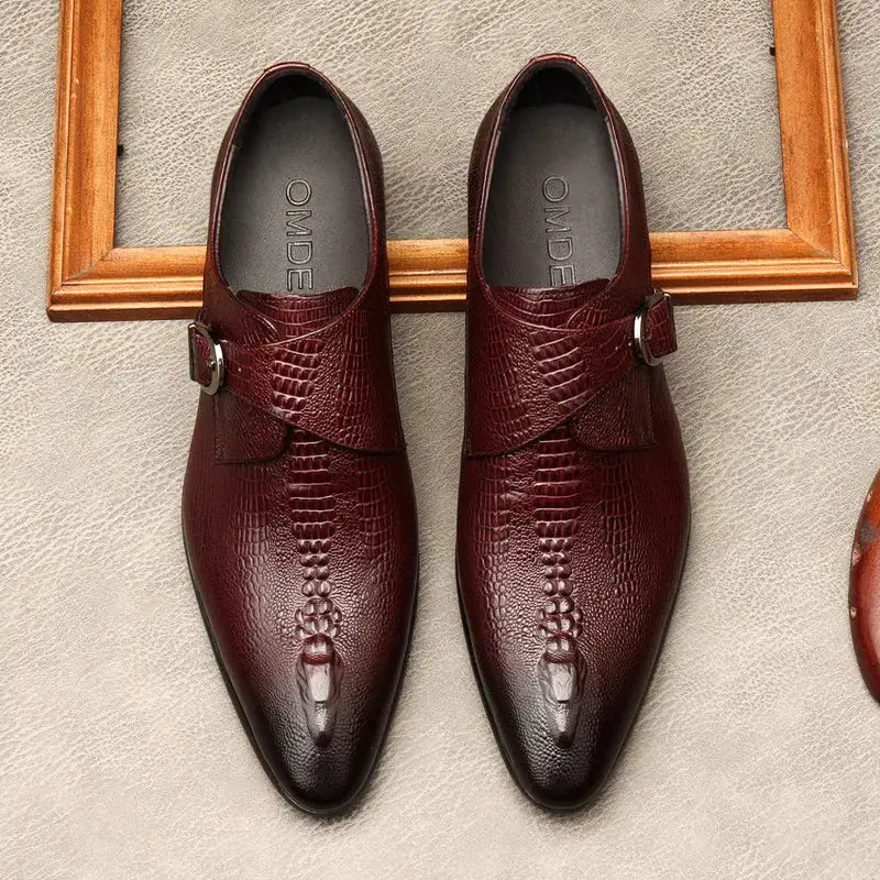 A pair of elegant burgundy leather loafers with a glossy finish. The shoes feature gold-toned metal horsebit detailing across the uppers and are displayed with wooden shoe trees inserted. They have a sleek, elongated silhouette with subtle stitching along the toe cap. The loafers are placed on a dark surface, partially framed by a wooden picture frame, emphasizing their classic and sophisticated style.