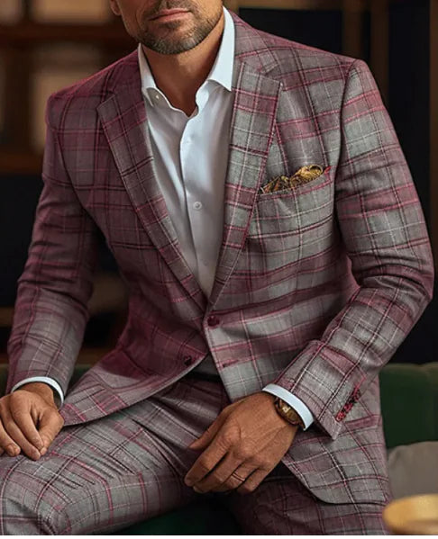 A stylish man wearing a tailored plaid suit in shades of gray and burgundy, paired with a crisp white dress shirt. The blazer features a subtle checkered pattern, and a patterned pocket square is neatly tucked into the breast pocket. He is accessorized with a brown leather-strap wristwatch. The background is softly blurred, showing a warm and sophisticated indoor setting.