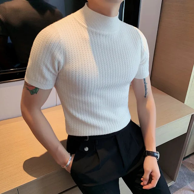 A man is wearing a stylish white short-sleeved turtleneck sweater with a textured knit pattern. The snug fit highlights his physique, and he pairs the sweater with tailored black trousers, creating a modern and sophisticated look. His accessories include a black wristwatch and a silver bracelet. The background features a sleek interior with a wooden surface and a mounted television, giving a refined, contemporary aesthetic.