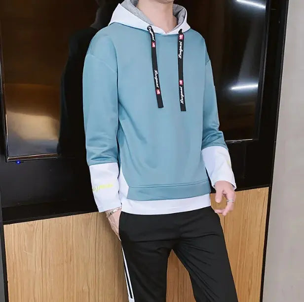 A stylish young man leans casually against a wooden wall while wearing a modern hoodie with black accents. The hoodie features black drawstrings with white text, black cuffs with embroidered details, and a red fabric tag on the left sleeve with 'LONG MA' printed on it. He is also wearing black athletic pants with white stripes on the sides. His accessories include silver rings on his fingers. The background consists of a contemporary indoor setting with a mix of wood and concrete elements.