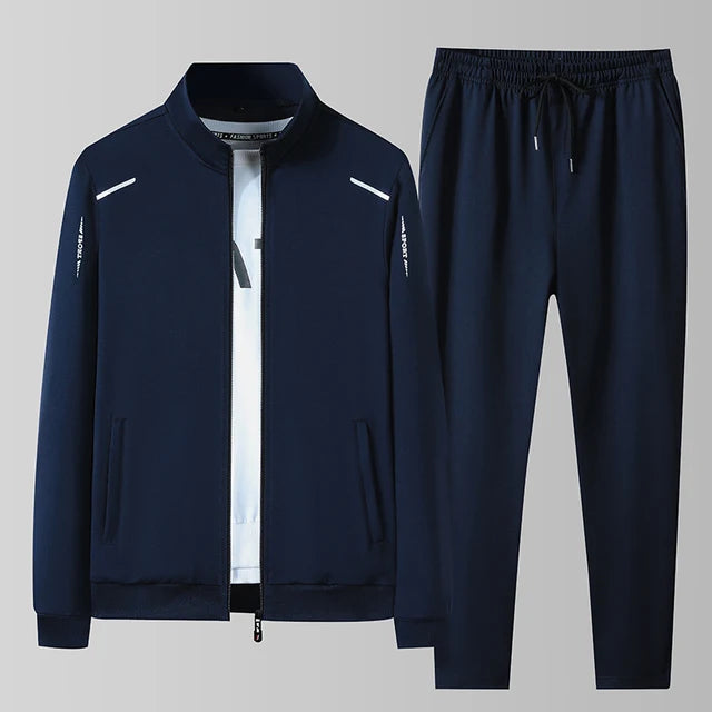 A navy blue athletic tracksuit consisting of a zip-up jacket and matching jogger pants, displayed against a light gray background. The jacket features a stand-up collar, full-length zipper, two side pockets, and subtle white accents on the shoulders and sleeves. The matching joggers have an elastic waistband with a drawstring, tapered legs, and side pockets. The fabric appears to be smooth and stretchy, suitable for sports or casual wear.