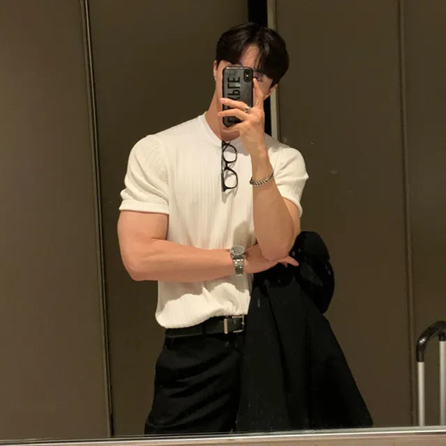 A stylish man taking a mirror selfie in a dimly lit modern space. He is wearing a fitted white short-sleeve shirt that highlights his toned arms, paired with black trousers and a belt. A black coat is draped over his arm, and a wristwatch and bracelet adorn his wrist. He holds his phone up to his face, partially covering it, and a pair of glasses hangs from his shirt collar. The background consists of sleek, reflective surfaces, adding a sophisticated touch to the scene.