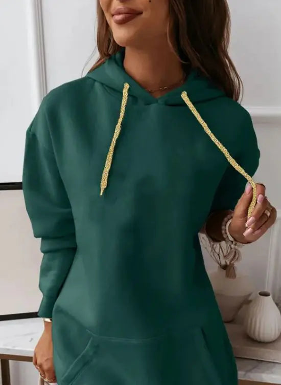 Woman wearing a long-sleeve, oversized maroon hoodie dress with a front kangaroo pocket and gold drawstrings. She is holding a light-colored handbag and posing indoors with a soft smile. The outfit has a casual, cozy look perfect for lounging or casual outings.