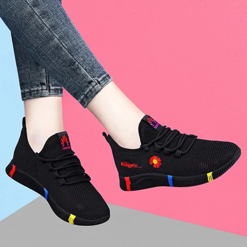 A pair of stylish black mesh sneakers worn with jeans. The shoes feature colorful embroidered details including a red and yellow daisy on the side and multicolored accents (red, blue, yellow) on the sole. The tongue displays a purple and red logo with the word "FASHION." The sneakers have a sleek, breathable design and a casual, trendy aesthetic.
