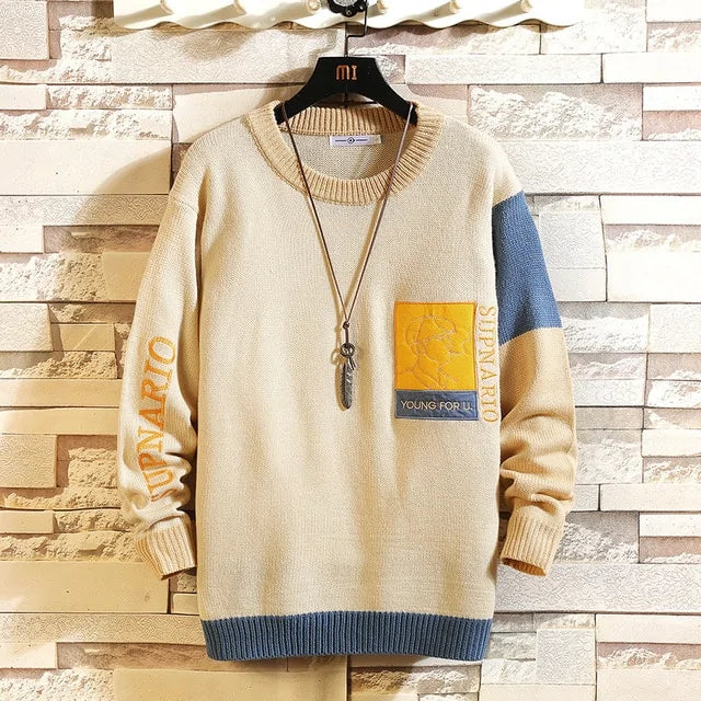 A fashionable knitted sweater with a beige body, blue ribbed hem, and beige ribbed cuffs and neckline. The left chest area features a yellow and blue patch with embroidered details, including the text "YOUNG FOR U." and "SUPNARIO." The left sleeve also has "SUPNARIO" embroidered in orange. One sleeve has a blue color block near the shoulder, while the other remains beige. The sweater is displayed on a black hanger against a textured brick wall background, with a long necklace featuring a feather pendant...