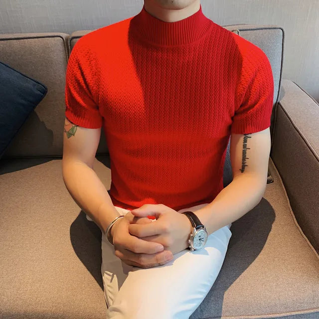 A man is wearing a bold red short-sleeved turtleneck sweater with a textured knit pattern. The sweater fits snugly, highlighting his physique. He pairs it with stylish white trousers, creating a sharp contrast. His accessories include a silver wristwatch and a bracelet. He is seated on a modern gray couch with a dark blue cushion in the background, giving a sophisticated and contemporary vibe.