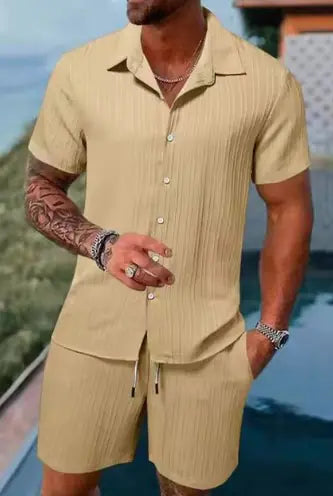 A stylish men's summer outfit featuring a short-sleeved, button-up shirt and matching shorts in a light beige color with a subtle vertical textured pattern. The outfit has a relaxed fit, ideal for warm weather. The man wearing it has a muscular build, accessorized with silver jewelry, including rings, a bracelet, and a chain necklace. His arms are tattooed, and he is standing near a poolside with a scenic outdoor background.