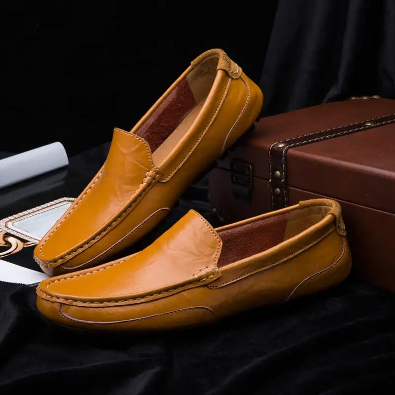 A pair of elegant burgundy leather loafers with a glossy finish. The shoes feature gold-toned metal horsebit detailing across the uppers and are displayed with wooden shoe trees inserted. They have a sleek, elongated silhouette with subtle stitching along the toe cap. The loafers are placed on a dark surface, partially framed by a wooden picture frame, emphasizing their classic and sophisticated style.