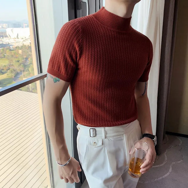 A man is wearing a rust-colored short-sleeved turtleneck sweater with a textured knit pattern. The fitted top pairs with high-waisted white trousers featuring a belt loop detail. He accessorizes with a silver bracelet, a black wristwatch, and holds a glass of amber-colored liquid. The setting is a modern high-rise apartment with a floor-to-ceiling window that offers a scenic view of a lush green landscape and city buildings in the distance. Natural light highlights the outfit's rich tones.
