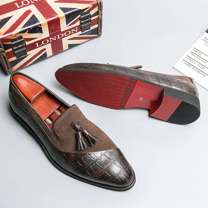 A pair of elegant burgundy leather loafers with a glossy finish. The shoes feature gold-toned metal horsebit detailing across the uppers and are displayed with wooden shoe trees inserted. They have a sleek, elongated silhouette with subtle stitching along the toe cap. The loafers are placed on a dark surface, partially framed by a wooden picture frame, emphasizing their classic and sophisticated style.