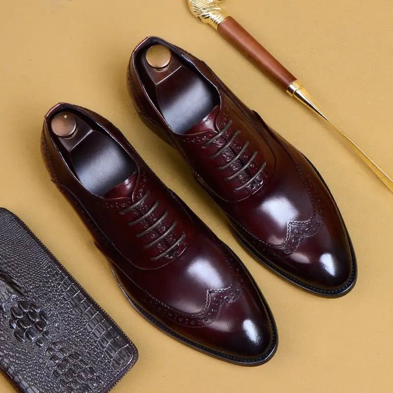 A pair of elegant burgundy leather loafers with a glossy finish. The shoes feature gold-toned metal horsebit detailing across the uppers and are displayed with wooden shoe trees inserted. They have a sleek, elongated silhouette with subtle stitching along the toe cap. The loafers are placed on a dark surface, partially framed by a wooden picture frame, emphasizing their classic and sophisticated style.