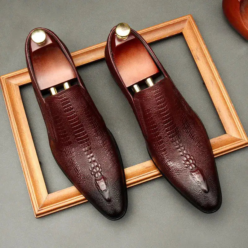 A pair of elegant burgundy leather loafers with a glossy finish. The shoes feature gold-toned metal horsebit detailing across the uppers and are displayed with wooden shoe trees inserted. They have a sleek, elongated silhouette with subtle stitching along the toe cap. The loafers are placed on a dark surface, partially framed by a wooden picture frame, emphasizing their classic and sophisticated style.