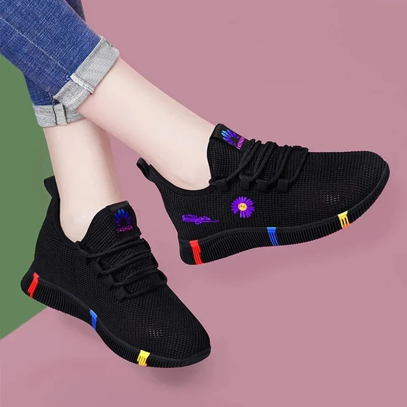 A pair of stylish black mesh sneakers worn with jeans. The shoes feature colorful embroidered details including a red and yellow daisy on the side and multicolored accents (red, blue, yellow) on the sole. The tongue displays a purple and red logo with the word "FASHION." The sneakers have a sleek, breathable design and a casual, trendy aesthetic.