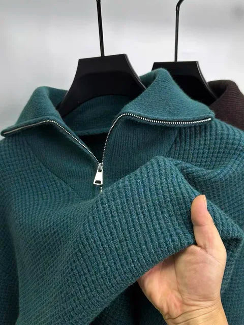 This image shows a close-up of a textured, waffle-knit pullover sweater in a rich burgundy color. It features a silver half-zipper with a folded-down collar, giving it a stylish and cozy look. A person is holding up the sleeve to showcase the fabric's softness and texture. In the background, another sweater in a teal shade is visible on a hanger, adding a contrast to the scene.