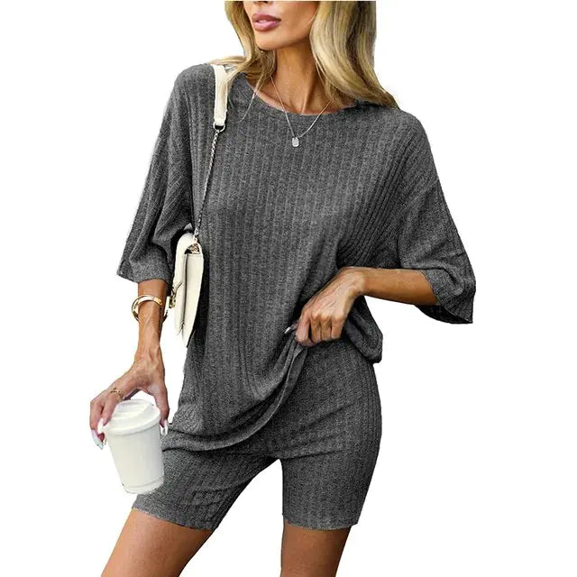 Woman wearing a matching ribbed mauve loungewear set consisting of a loose-fitting oversized top and fitted shorts, accessorized with a crossbody bag, bracelet, and holding a white takeaway coffee cup.