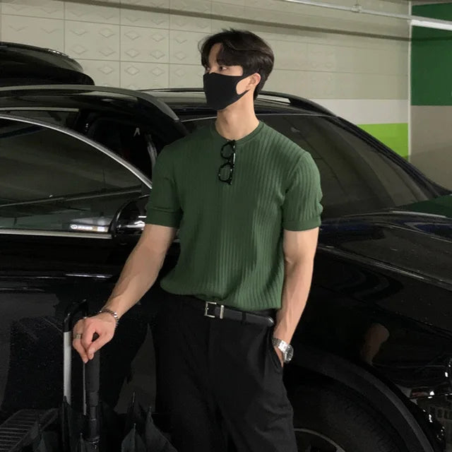 A stylish man with a fit physique is seen leaning against a black luxury car in an indoor parking garage. He is wearing a fitted green ribbed short-sleeve shirt tucked into black trousers with a sleek black belt. A pair of glasses hangs from the collar of his shirt. He accessorizes with a silver wristwatch and a bracelet on his left wrist. His face is partially covered by a black mask, and his dark hair is neatly styled. His posture is relaxed, with one hand resting on an umbrella handle while gazing off...