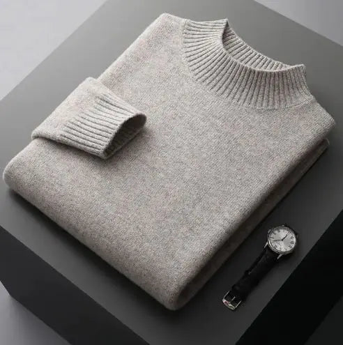 A neatly folded beige knitted sweater with a ribbed high collar and cuffs, placed on a dark grey surface. Beside the sweater, a classic wristwatch with a black leather strap and a silver dial is positioned, adding a touch of elegance to the composition.