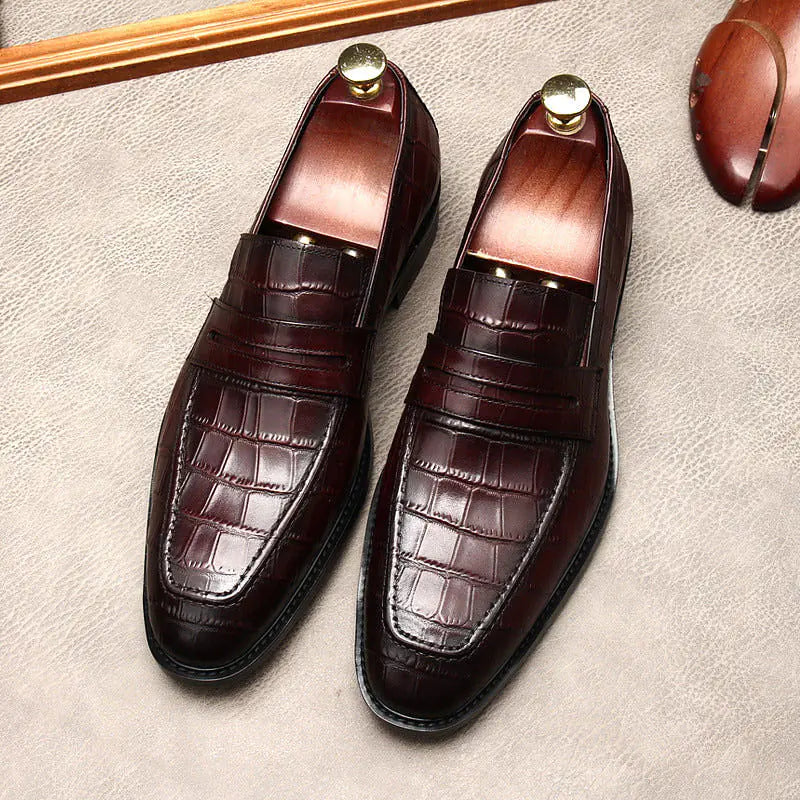 A pair of elegant burgundy leather loafers with a glossy finish. The shoes feature gold-toned metal horsebit detailing across the uppers and are displayed with wooden shoe trees inserted. They have a sleek, elongated silhouette with subtle stitching along the toe cap. The loafers are placed on a dark surface, partially framed by a wooden picture frame, emphasizing their classic and sophisticated style.