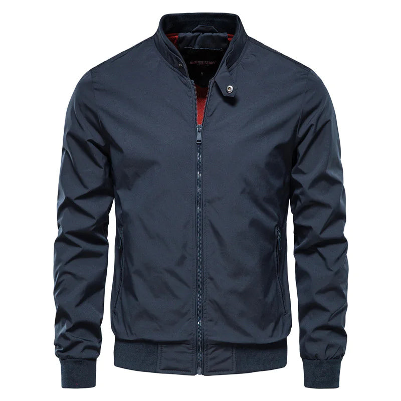 A sleek navy blue bomber jacket with a zip-up front, ribbed cuffs, and hem. The jacket features a high collar with a snap button closure and a subtle red inner lining, adding a touch of contrast. It has a modern, slim-fit design with two zippered side pockets, making it a stylish and functional outerwear choice.