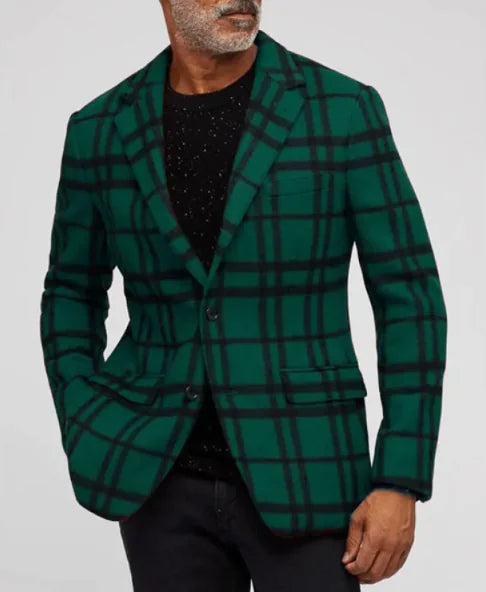A stylish man with a well-groomed beard wears a green plaid blazer with black intersecting lines. The blazer features a notched lapel, two-button closure, and flap pockets. Underneath, he sports a black speckled shirt, paired with dark-colored pants. His left hand is casually placed in his pocket, and his right hand hangs naturally by his side. The background is a simple, neutral gray, highlighting the bold pattern of the blazer.