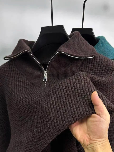 This image shows a close-up of a textured, waffle-knit pullover sweater in a rich burgundy color. It features a silver half-zipper with a folded-down collar, giving it a stylish and cozy look. A person is holding up the sleeve to showcase the fabric's softness and texture. In the background, another sweater in a teal shade is visible on a hanger, adding a contrast to the scene.