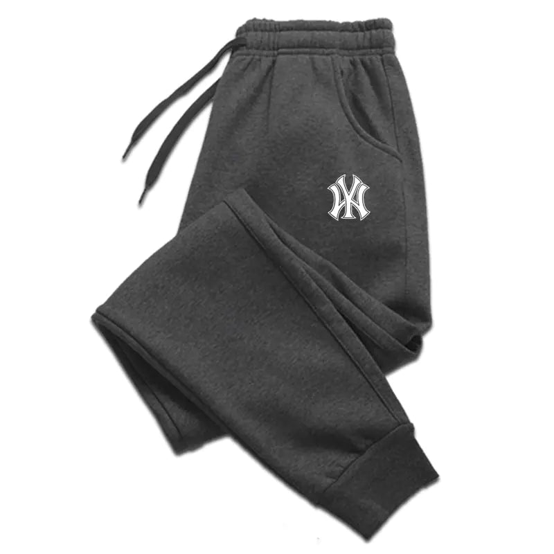 A pair of dark gray sweatpants with an elastic waistband and drawstrings, laid flat against a white background. The sweatpants feature ribbed cuffs at the ankles and side pockets. A black embroidered logo, resembling the New York Yankees emblem, is visible on the upper left thigh. The fabric appears to be soft, suggesting a comfortable and relaxed fit.