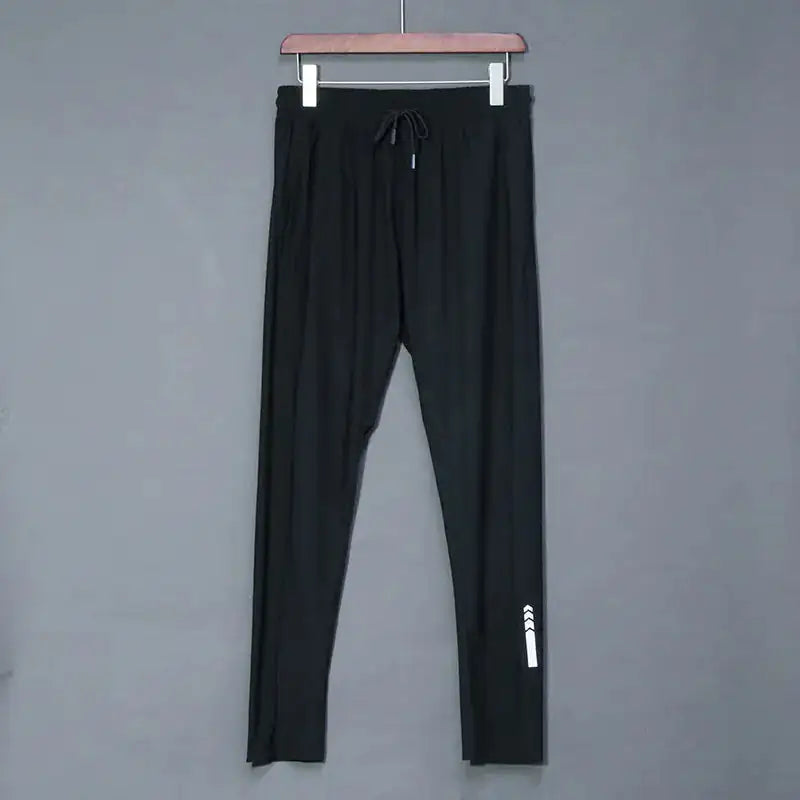 A pair of black athletic joggers hangs on a wooden hanger against a plain gray background. The joggers feature an elastic waistband with a drawstring for adjustable fit. A small white reflective design is visible near the lower leg, adding a subtle sporty touch. The fabric appears lightweight and stretchy, making the joggers suitable for casual wear, workouts, or lounging.