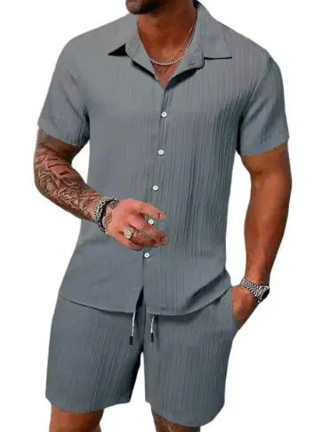A stylish man with a muscular build is wearing a short-sleeved, button-up green shirt with matching green shorts. He is accessorized with silver rings, a wristwatch, and a layered necklace. His left arm is tattooed, and he is posing casually near a modern poolside setting with a blurred outdoor background.