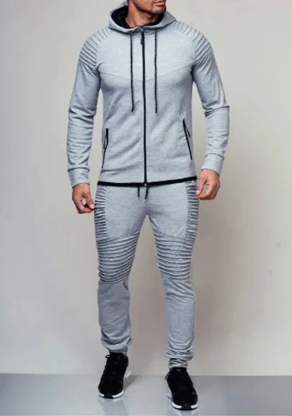 A man is wearing a stylish light gray tracksuit with a fitted design. The zip-up hoodie features ribbed detailing on the shoulders, black drawstrings, and zippered pockets. The matching joggers have ribbed accents on the thighs and knees, along with black drawstrings at the waist. He is also wearing black sneakers with white soles. The neutral background enhances the modern and sporty look of the outfit.
