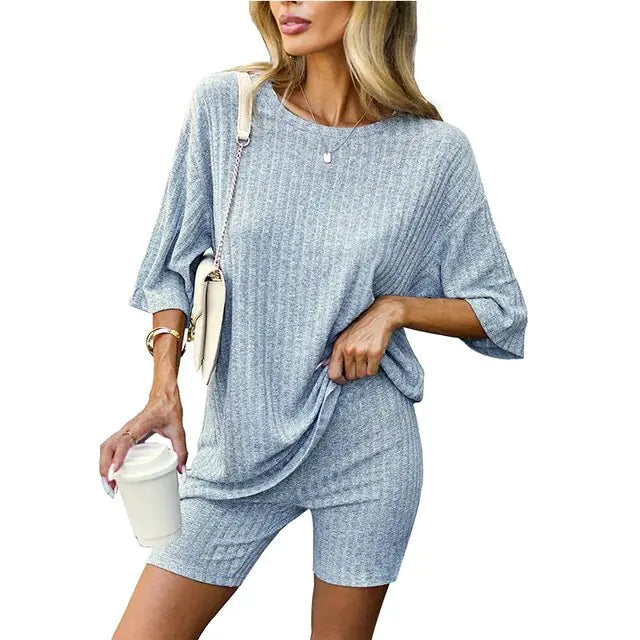 Woman wearing a matching ribbed mauve loungewear set consisting of a loose-fitting oversized top and fitted shorts, accessorized with a crossbody bag, bracelet, and holding a white takeaway coffee cup.