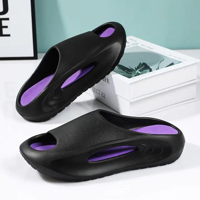 A pair of stylish black slip-on sandals with purple cushioned insoles, featuring cutout designs on the sides and toes, displayed on a white surface next to stacked books.