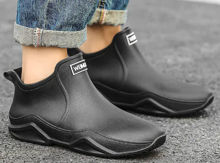A pair of black waterproof ankle rain boots worn with cuffed jeans. The boots have a sleek, modern design with thick, textured soles and a branded label on the front that reads "WEMAM." The setting is an outdoor pavement, suggesting the boots are suited for wet or rainy weather.