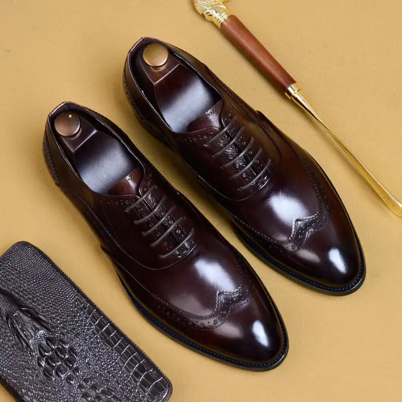 A pair of elegant burgundy leather loafers with a glossy finish. The shoes feature gold-toned metal horsebit detailing across the uppers and are displayed with wooden shoe trees inserted. They have a sleek, elongated silhouette with subtle stitching along the toe cap. The loafers are placed on a dark surface, partially framed by a wooden picture frame, emphasizing their classic and sophisticated style.