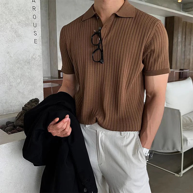 A fashionable man wearing a fitted, ribbed brown polo shirt with short sleeves and a buttoned collar. A pair of black-framed eyeglasses hangs from the collar. He is also wearing light-coloured trousers and a silver wristwatch. In one hand, he holds a black blazer. The background features a modern, neutral-toned interior with lounge seating and decorative stone elements.