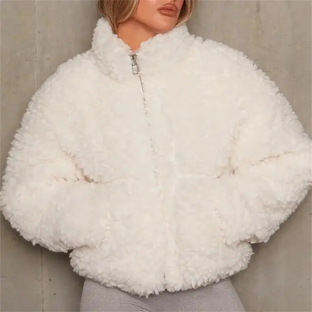 Woman wearing a white, fluffy sherpa-style zip-up jacket with a high collar and side pockets. The cozy jacket has a soft, textured appearance and is styled with light-colored pants, photographed against a neutral concrete background.