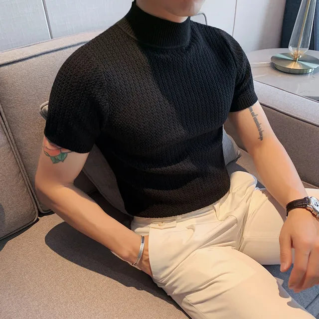 A man is seated on a modern gray sofa, wearing a black short-sleeved turtleneck sweater with a textured knit pattern. The snug fit of the sweater accentuates his physique. He pairs it with high-waisted cream trousers, secured with a belt. His accessories include a silver bracelet on his left wrist and a black wristwatch on his right. His arms, which have visible tattoos, rest casually on the couch. The background features a contemporary interior with a side table, a sleek lamp, and neutral-colored...