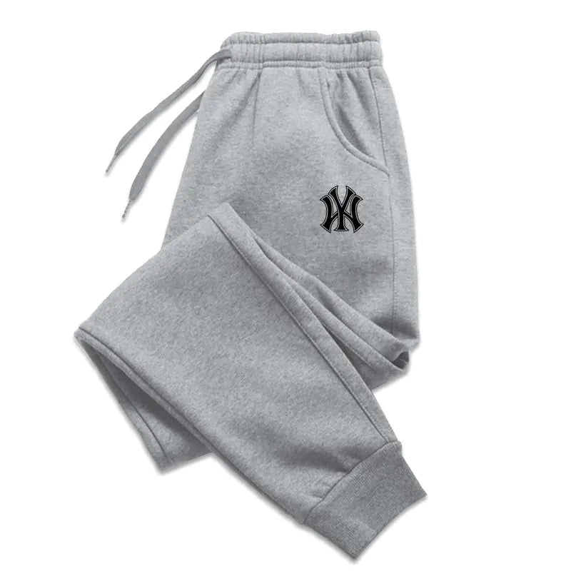 A pair of light gray sweatpants with an elastic waistband and drawstrings, laid flat against a white background. The sweatpants feature ribbed cuffs at the ankles and side pockets. A black embroidered logo, resembling the New York Yankees emblem, is visible on the upper left thigh. The fabric appears to be soft, suggesting a comfortable and relaxed fit.